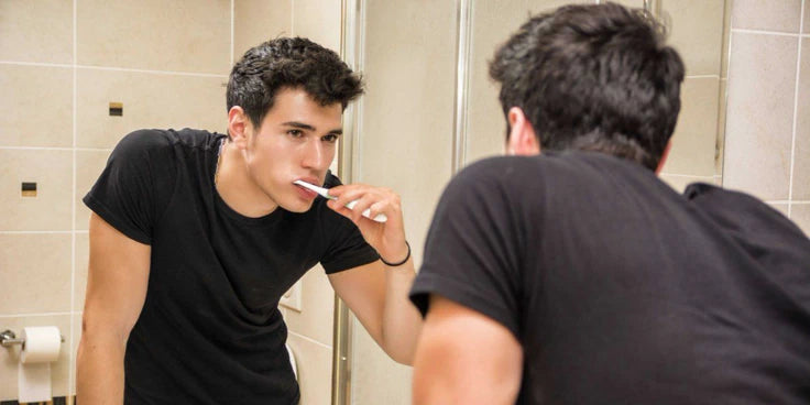 A young man in a black t-shirt brushing his teeth in a tiled bathroom, seen both in the mirror and from behind.