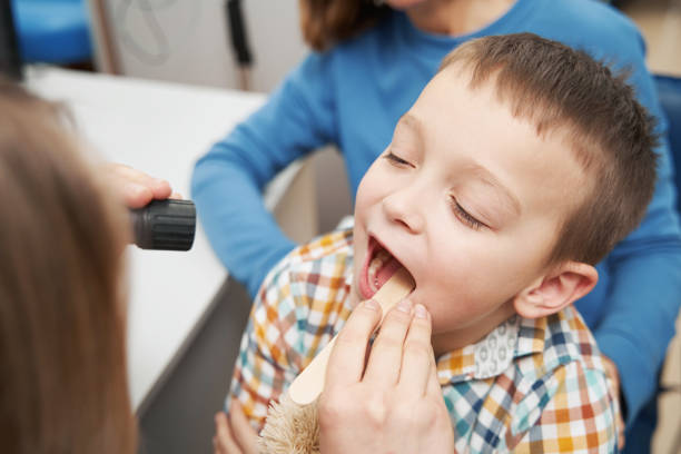 A young boy with his mouth open receives a dental exam or treatment, with a dentist holding a light source and another person in a blue shirt behind him.