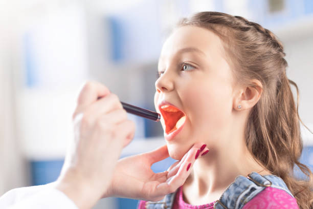 A close-up lifestyle shot of a young girl with her mouth open as a dentist or hygienist examines her teeth with a dental mirror. The girl is wearing a denim vest over a pink top, and the background is a brightly lit, blurred clinic interior.